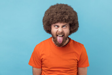 Fototapeta na wymiar Portrait of man with Afro hairstyle wearing orange T-shirt showing out tongue and crossed eyes with naughty disobedient grimace, making face. Indoor studio shot isolated on blue background.