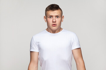 Fototapeta premium Portrait of astonished surprised attractive teenager boy wearing T-shirt standing looking at camera with big eyes, being frighten and scared. Indoor studio shot isolated on gray background.