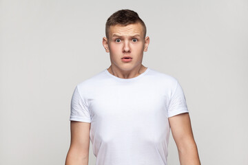 Portrait of astonished surprised attractive teenager boy wearing T-shirt standing looking at camera with big eyes, being frighten and scared. Indoor studio shot isolated on gray background.