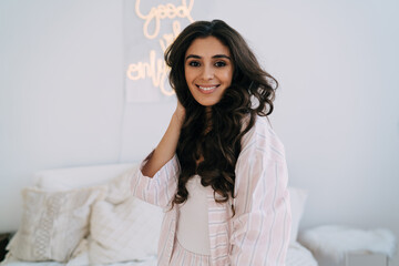 Naklejka premium Happy ethnic lady smiling and touching wavy hair in bedroom