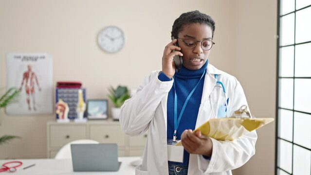 African american woman doctor reading document on clipboard talking on smartphone at clinic