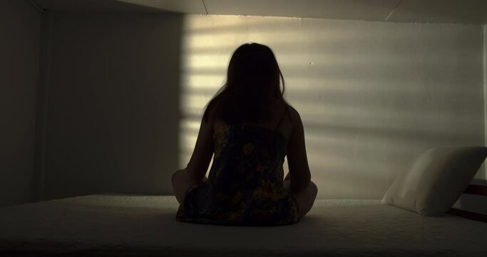 A lonely woman is sitting on the bed in a narrow dark room. This concept use for symptoms of stress, major depressive disorder (MDD), psychotic symptoms.