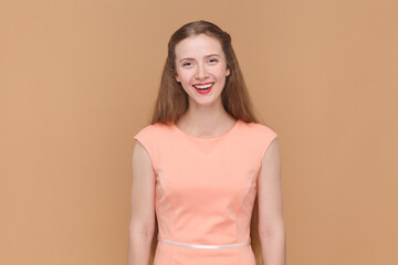 Obraz premium Portrait of joyful cheerful beautiful woman with long hair looking at camera with toothy smile, being in good mood, wearing elegant dress. Indoor studio shot isolated on brown background.