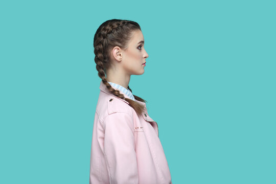 Side view portrait of strict bossy young teenager girl with braids wearing pink jacket looking ahead with serious facial expression, being in bad mood. Indoor studio shot isolated on green background.
