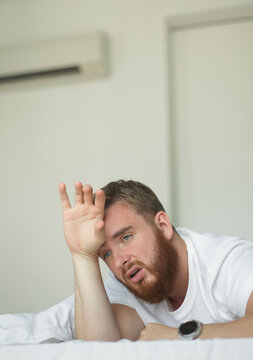 Young bearded man using air conditioner at home, lay on bed cooling off during hot weather, suffering from heat, high temperature. Broken AC. Vertical photo. 