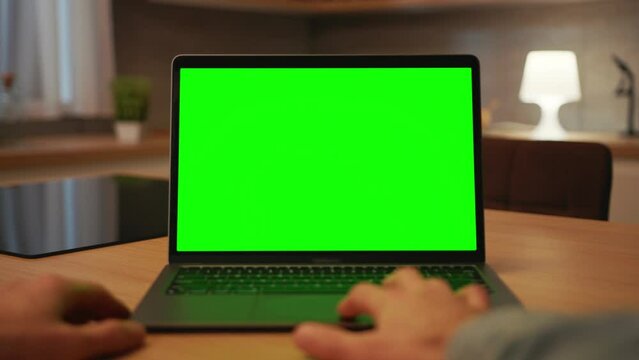 Footage of computer green screen. Shot of unrecognizable Caucasian man typing on keyboard of laptop in blurred view of kitchen in evening. Horizontal shot of male hands. Remote work. Indoors