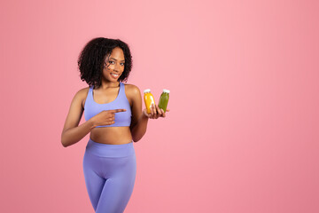 Positive millennial black lady in sportswear hold jars of juice or smoothies isolated on pink...