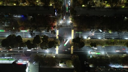 It's raining in the city. View of the intersection. There are green buses, there are cars. Lights from vehicles, lanterns and houses are reflected on asphalt. View from above. There are green trees
