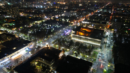 A big concert in the night city. Drone view of a crowd of people, rays and lights of light. A large building with lighting. There are roads with cars, pedestrian streets with people and a park around