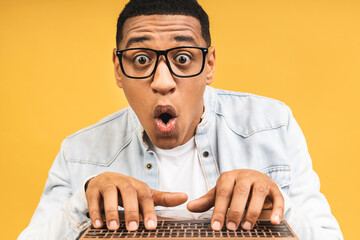 Young smiling shocked amazed african american man standing and using laptop computer isolated over yellow background.
