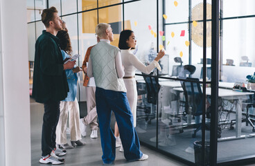 Group of experienced colleagues discussing information from modern glass board with memo sticks, professional business partners collaborating on startup and business project talking in workspace