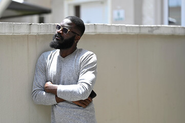 Happy young man standing with arms crossed against a wall and looking away, with sunglasses on 