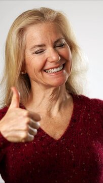 Vertical video giving a thumbs up, a pretty attractive mature blond woman in red sweater smiling happy portrait isolated on solid white background with copy space.