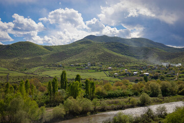 A beautiful rural panorama located under mountains, near the river reflecting bright sunlight, on a partially cloudy spring day