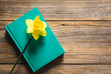 Green book and yellow narcissus flower on wooden background, top view.