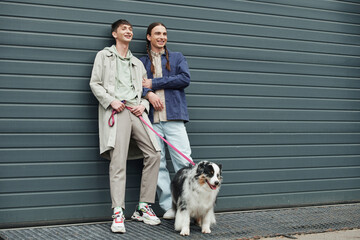 cheerful gay man holding leash of Australian shepherd dog and standing next to smiling boyfriend with pigtails near garage door outside on urban street - Powered by Adobe
