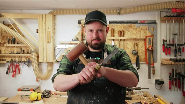 Caucasian young carpenter with a protective apron holding a mallet and a chisel with hands crossed and smiling surrounded by many tools. High quality 4k footage