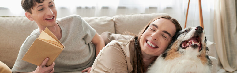 happy gay man with tattoo smiling while hugging Australian shepherd dog next to cheerful gay man holding book and resting on comfortable couch in living room, banner