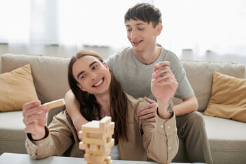 Smiling gay man hugging and looking at long haired and tattooed boyfriend while playing blurred wood blocks game on table near couch in living room at home