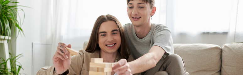 Young and smiling same sex couple in casual clothes playing blurred wood blocks game near couch and...