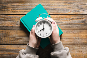 Alarm clock in female hands and a green book on a wooden background, top view.