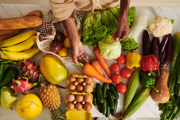 Fresh veggies, fruits, eggs and bread on kitchen table in front of Black woman