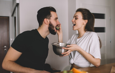 Beautiful Caucasian wife spoon-feed her husband enjoying time for eating togetherness during breakfast in home apartment, Carefree woman with salad plate giving vegetables to vegan boyfriend