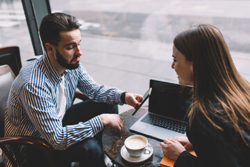 Cheerful couple using smartphone and laptop in modern cafeteria