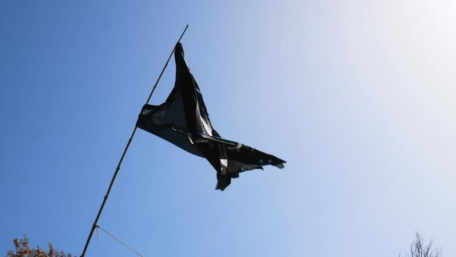 Completely torn and bruised black colored pirate flag (Jolly Roger) fastened to a flagpole flutters in the wind with a skull with patch and two swords on the bottom in white drawn on the fabric