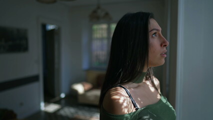 One contemplative young woman standing by window looking outside with pensive expression. Thoughtful female perso in 20s stands inside home