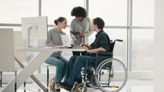 Young women and disabled man in wheelchair are talking and working together in office. Creative business and people concept. Multiracial business people working inside office.
