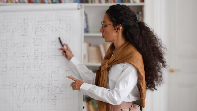 Professional young woman teacher in eyeglasses nurturing young minds online, explaining new math topic on whiteboard