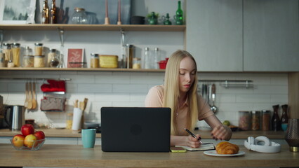 Girl student studying online sitting at kitchen with laptop. Woman making notes.
