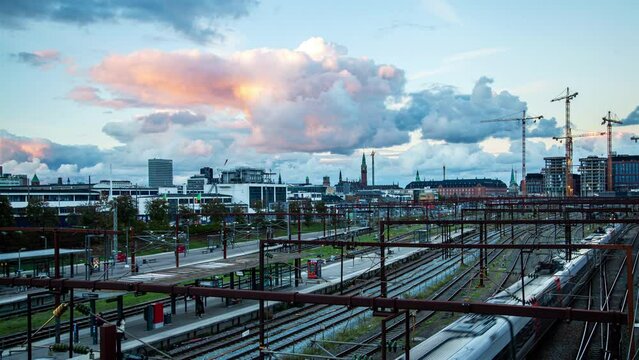 Timelapse of multi-lane railway tracks and urban architecture at Copenhagen's central station during autumn sunset, featuring a cloudy skyline.
