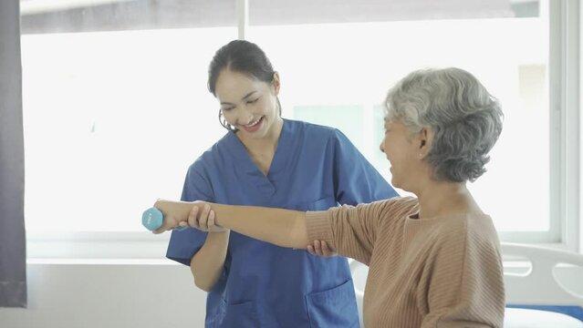 Young female physiotherapist talking and showing arm movements to gray-haired old woman using resistance bands for muscle exercise and physiotherapy