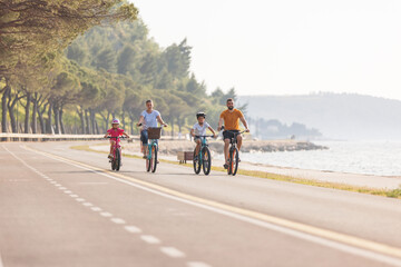 Obraz premium Two kids, with helmets on their heads, and smiling parents riding bikes on a family-friendly cycle route along a sea coastline, front view.
