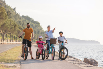 Young family, parents with two children, on bikes standing near the sea, looking at view in the direction that the mother pointing