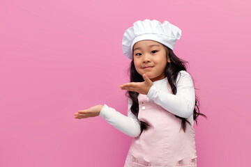little asian girl in chef uniform smiles and shows her hands to the side on pink isolated...