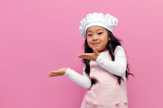 little asian girl in chef uniform smiles and shows her hands to the side on pink isolated background, korean child cook