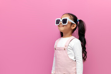 little girl of preschool age in white sunglasses and pink sundress on pink isolated background, child in glasses looks