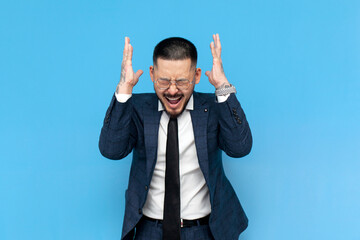 annoyed asian businessman in suit and glasses screaming in stress on blue isolated background,...