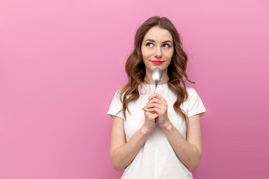 hungry woman licks spoon and dreams on pink isolated background, pensive girl in white t-shirt thinks about food