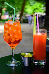 Aperol Sprits summer cocktail with Aperol, prosecco, ice cubes and orange in wine glass and glass of fresh made tomato juice ready to drink on sunny terrace