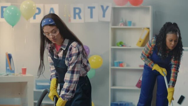 Asian woman and African American woman cleaning the office after a party. Cleaning ladies in blue overalls mop the floor and vacuum. The concept of cleaning and hygiene service.