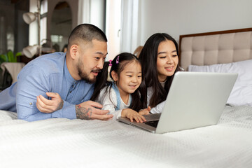 asian family use laptop at home on bed, korean dad mom and little daughter look at computer screen...