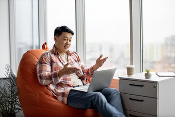 Positive asian worker in casual wear taking part in online briefing on laptop in spacious office with panoramic windows. Young man gesturing hands while explaining details of new business project.