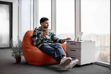 Young indian man in casual outfit and white sneakers sitting on orange bean bag chair and working...