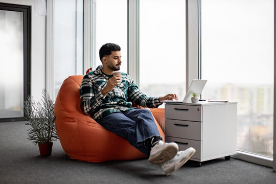 Young indian man in casual outfit and white sneakers sitting on orange bean bag chair and working on laptop in comfortable office. Focused worker holding cup of hot coffee and typing on keyboard.