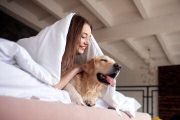 girl lies in bed under warm blanket with golden retriever dog and looks out the window in the morning, woman with pet