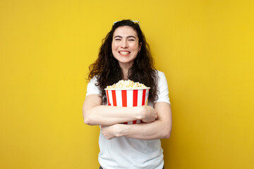 young curly cheerful girl viewer in white t-shirt holding popcorn on yellow background and smiling